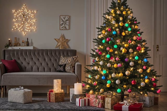 A Christmas tree with colorful lights and ornaments in front of a couch. The tree is surrounded by presents and is the centerpiece of the room.