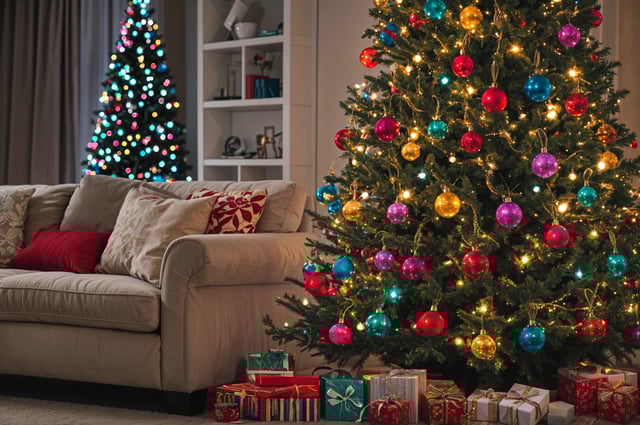 A Christmas tree with colorful ornaments and presents underneath it. The tree is in front of a couch and a bookshelf. The scene is decorated for the holiday season.